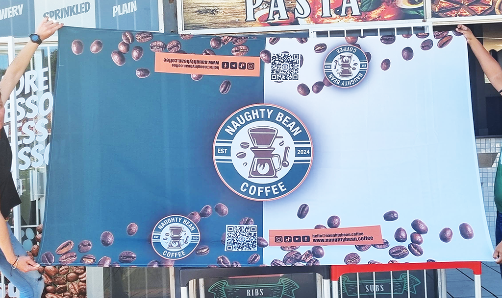 A branded tablecloth printed by Brand Monster is being held by two customers at Naughty Bean Coffee Shop. The tablecloth is printed with Naughty Bean's logo and artwork.